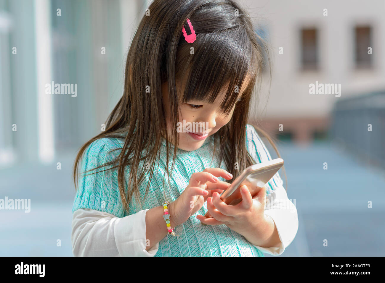 Piuttosto alla moda bambina gioca con un telefono cellulare all'aperto sotto il sole sorride mentre naviga sullo schermo Foto Stock