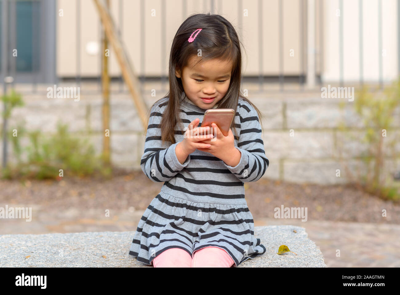 Carino bambina affascinato da un telefono cellulare seduti all'aperto la lettura dello schermo con una felice espressione Foto Stock