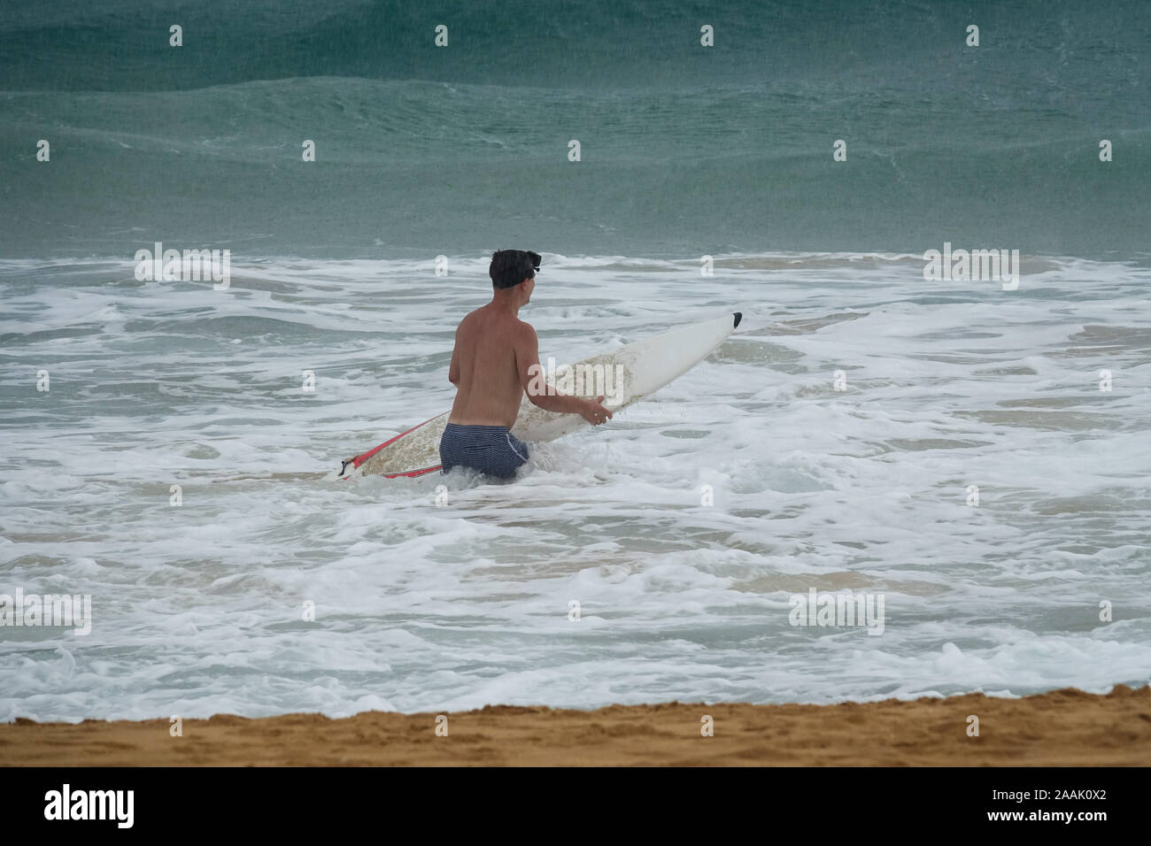 Giovane uomo con la tavola da surf in esecuzione nell'oceano sotto la pioggia Foto Stock
