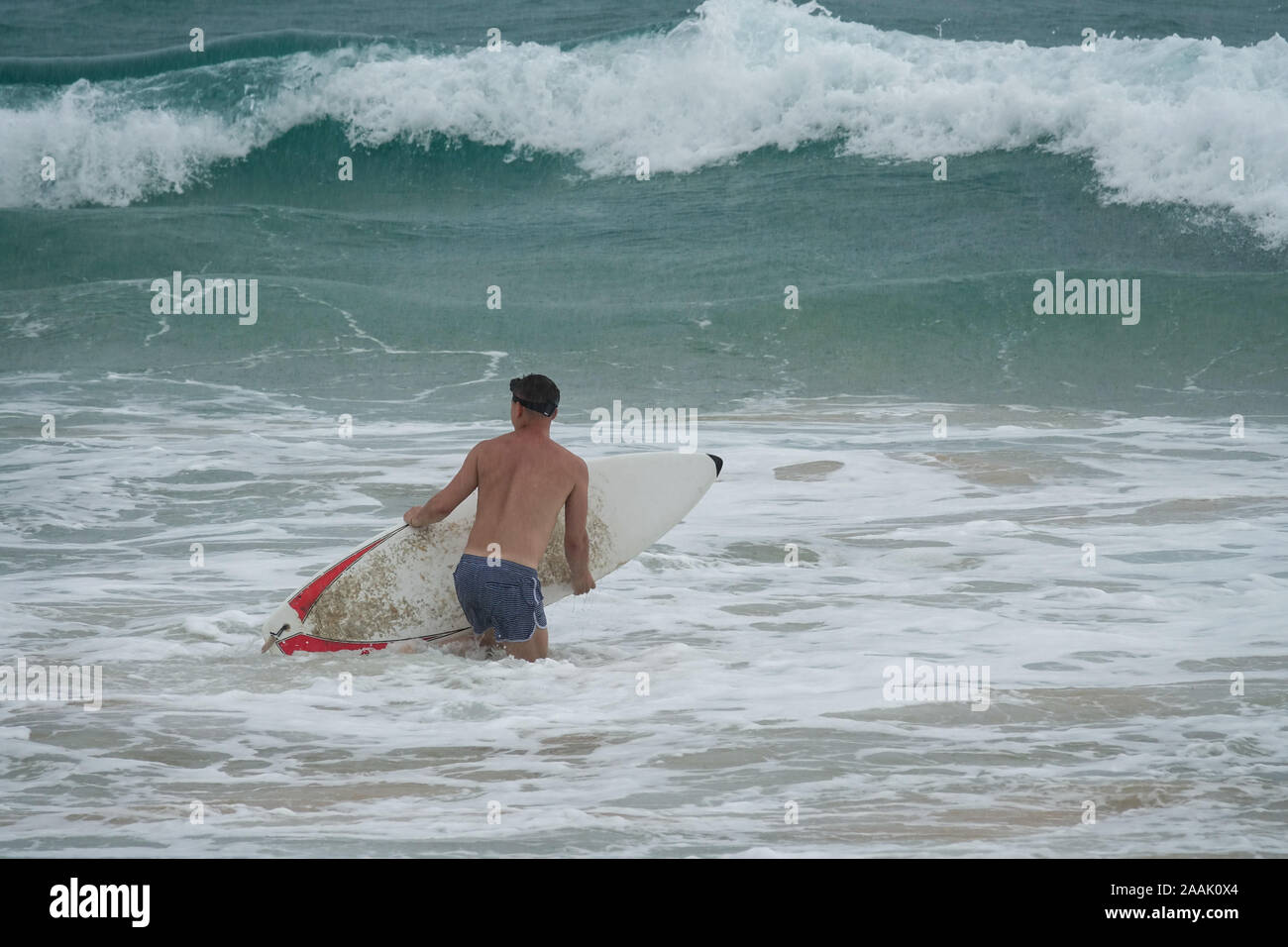 Giovane uomo con la tavola da surf in esecuzione nell'oceano sotto la pioggia Foto Stock
