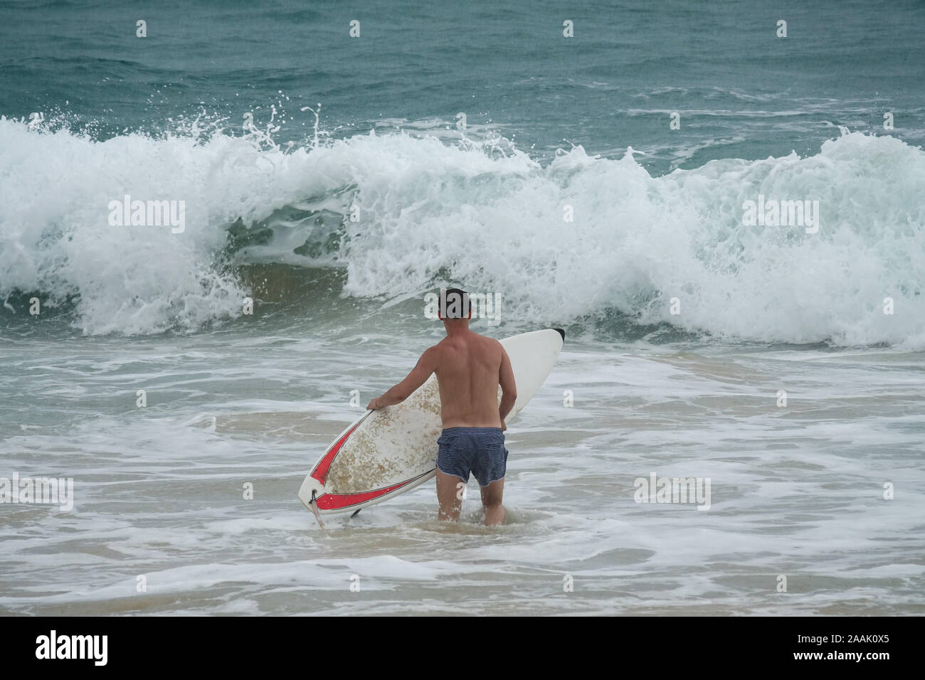Giovane uomo con la tavola da surf in esecuzione nell'oceano sotto la pioggia Foto Stock