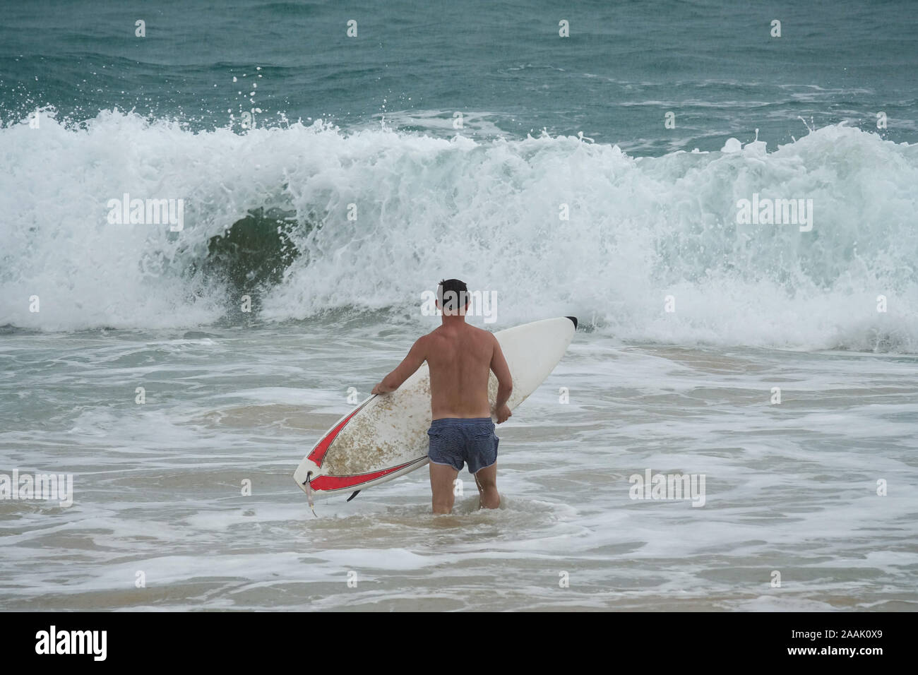 Giovane uomo con la tavola da surf in esecuzione nell'oceano sotto la pioggia Foto Stock