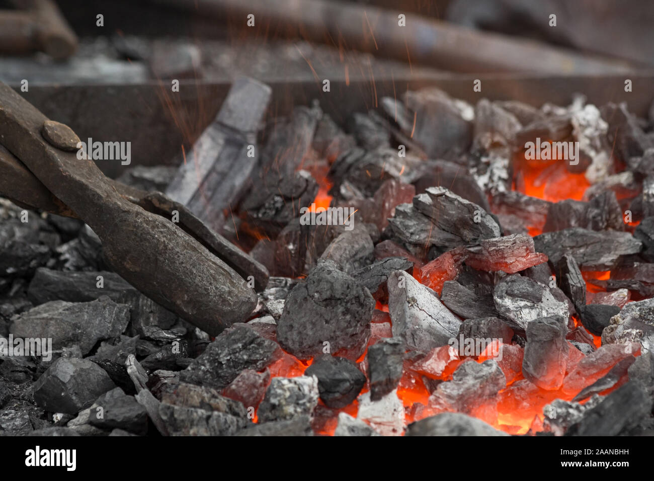 Un fabbro imposta il braciere con tizzi con grandi tenaglie di carbone. Embers glow in una fucina di ferro. Incendio, calore, carbone e cenere con scintille volanti. Foto Stock