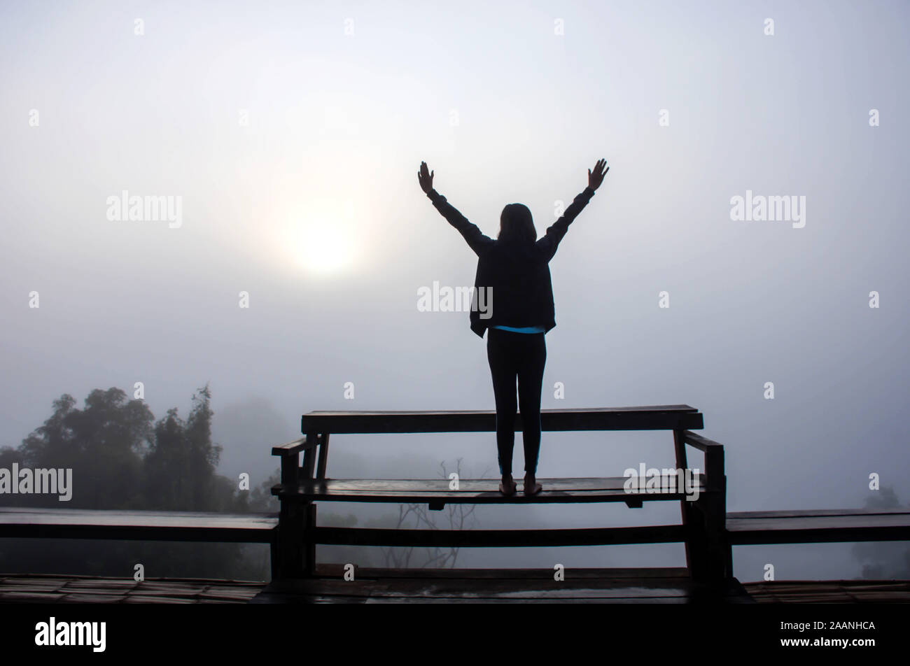 L'immagine dietro le donne sollevare le braccia su una sedia in legno sullo sfondo la nebbia e la luce del sole di mattina. Foto Stock