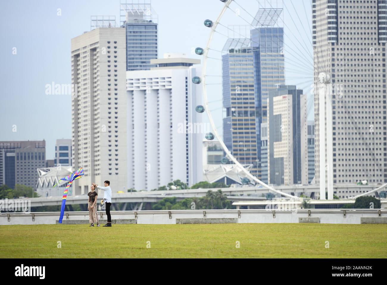 Una bella coppia felice stanno giocando con un aquilone in un parco pubblico con il traghetto a ruota e lo skyline di Singapore in background. Foto Stock