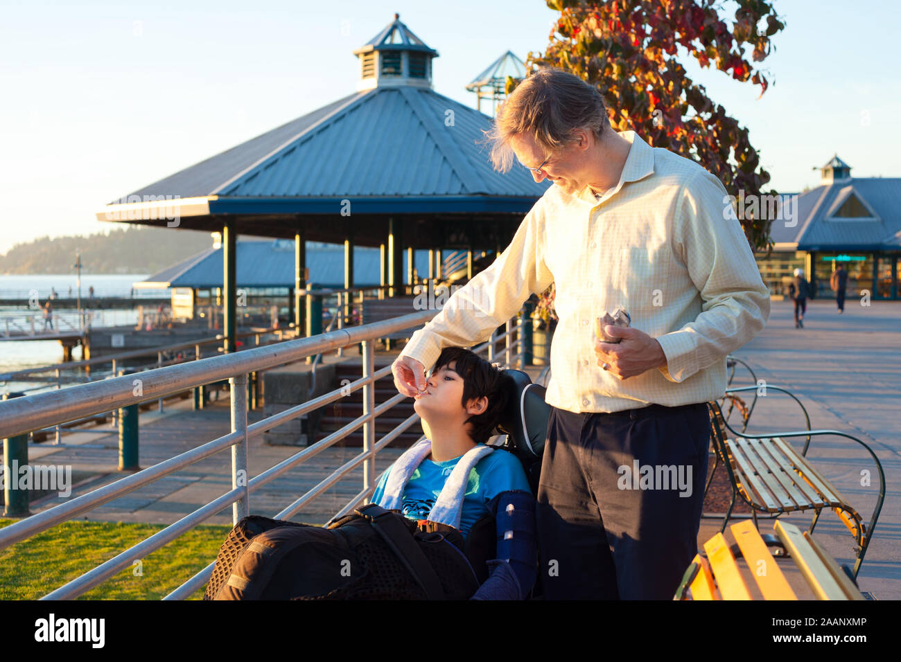 Padre caucasici in quaranta biracial alimentazione figlio disabili in sedia a rotelle al parco sulla giornata di sole Foto Stock