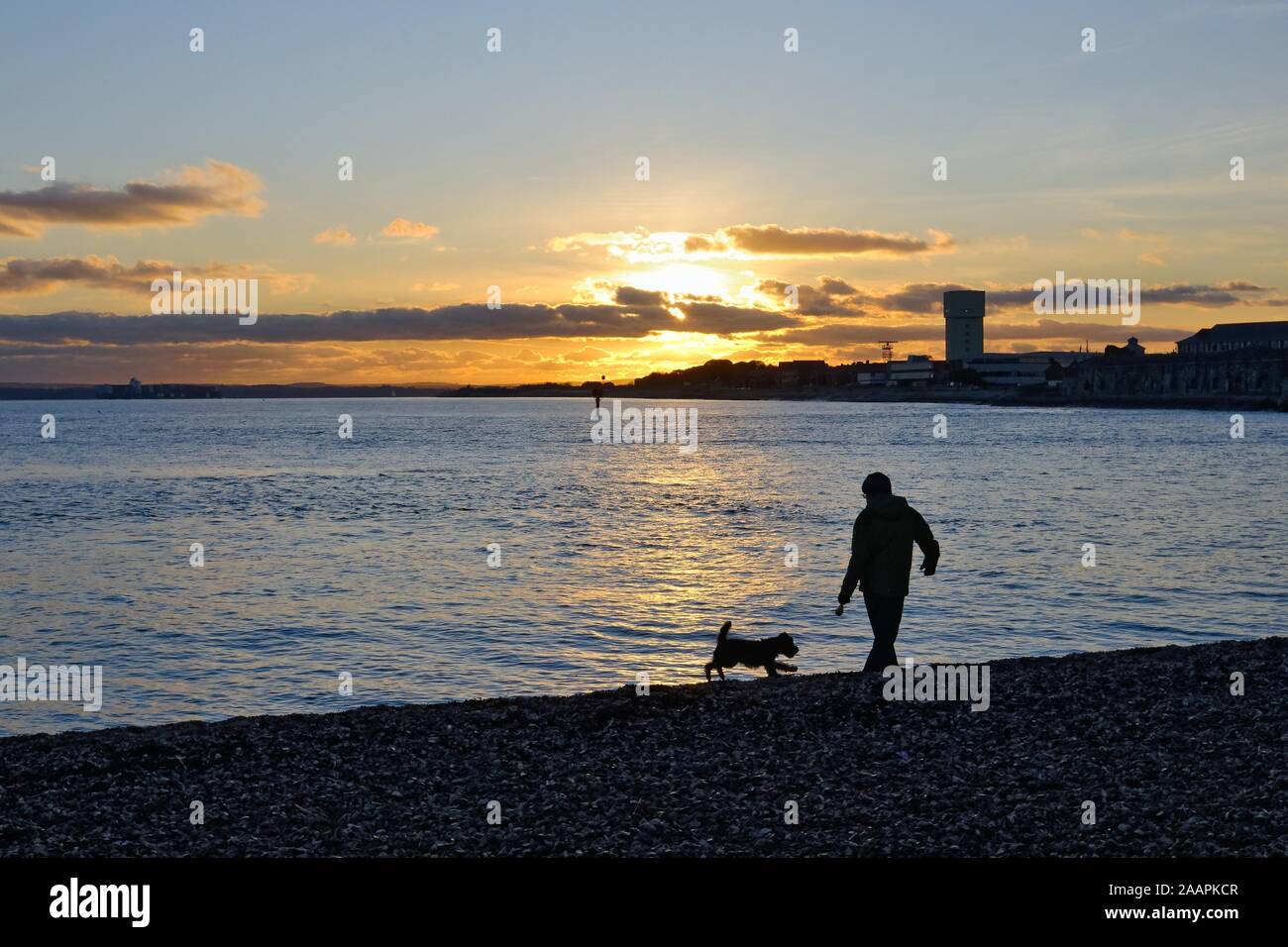 Silhouette di un uomo a piedi un cane sulla spiaggia a Portsmouth contro un tramonto in inverno, Hampshire England Regno Unito Foto Stock
