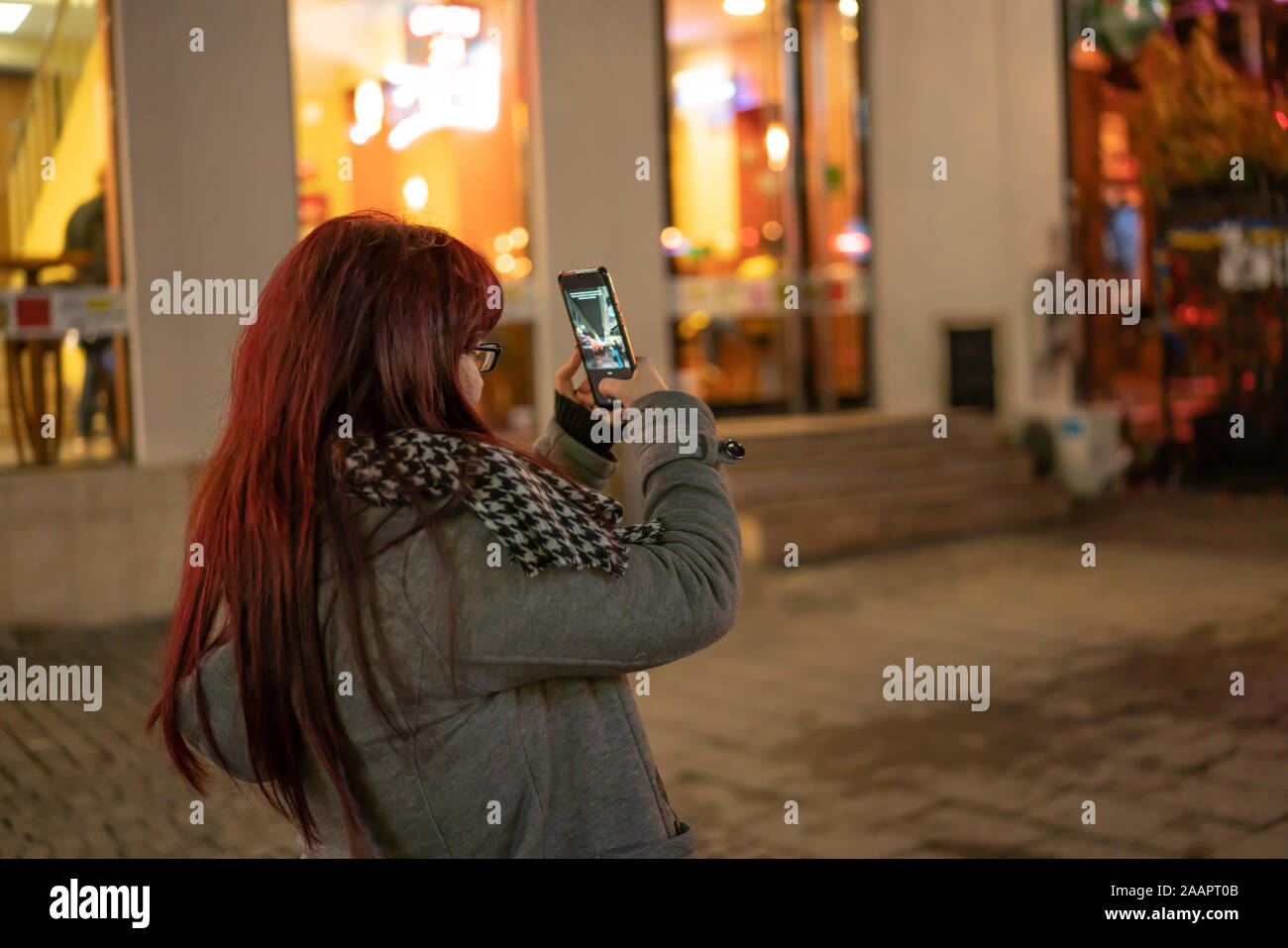 Bella e giovane europeo redhead ragazza con gli occhiali di scattare una foto con il suo smartphone di notte in una strada nel centro di Bucarest (Romania) Foto Stock