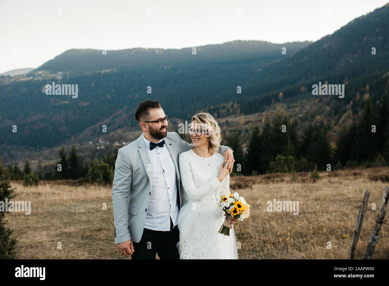 Bella giovane a piedi e sorridente sulle loro operazioni di diserbatura Giorno, nelle montagne al tramonto. Sposa è in un bianco abito da sposa con un bouquet di girasoli. Foto Stock