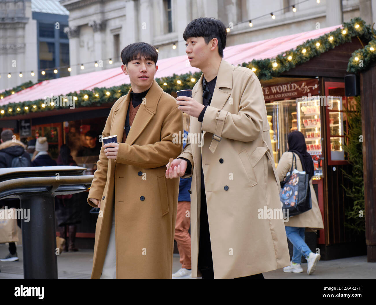 Due asian giovane trenchcoats bere cioccolata calda dal mercato di Natale di stallo a Trafalgar Square Foto Stock