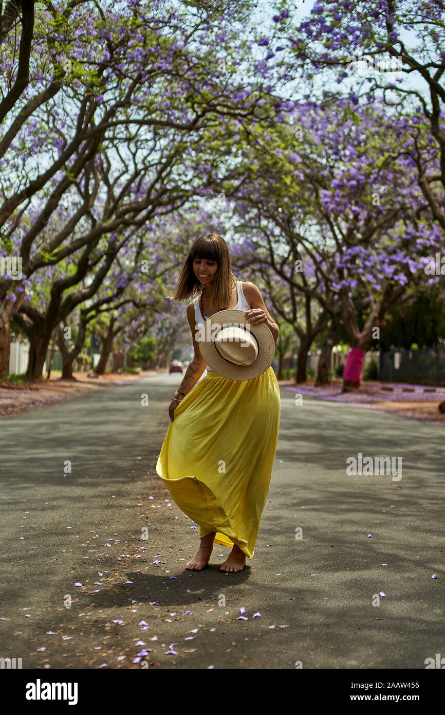 Woman Dancing con un cappello sulle sue mani nel mezzo di una strada piena di alberi di jacaranda in fiore Pretoria, Sud Africa Foto Stock