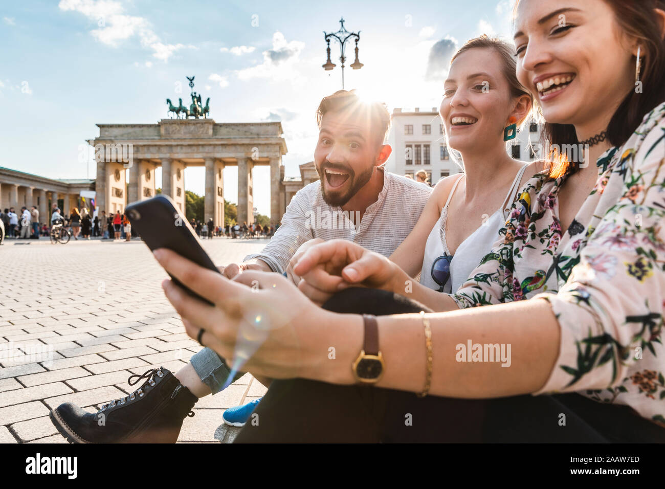 Tre ridere gli amici seduti vicino al Brandenburger Tor guardando smartphone, Berlino, Germania Foto Stock