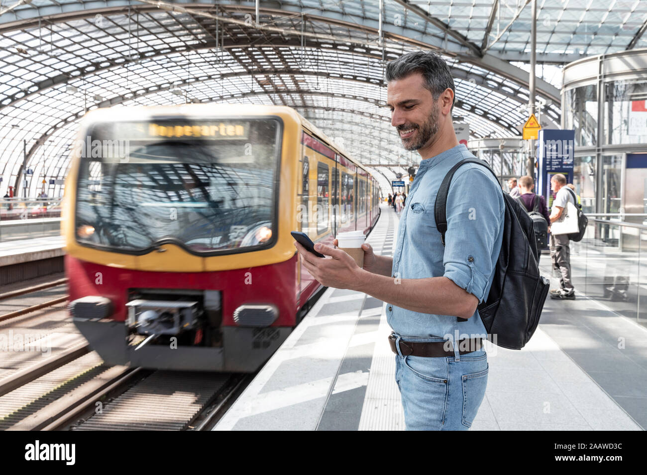 Un uomo d'affari sorridente alla stazione in attesa del treno che guarda lo smartphone, Berlino, Germania Foto Stock