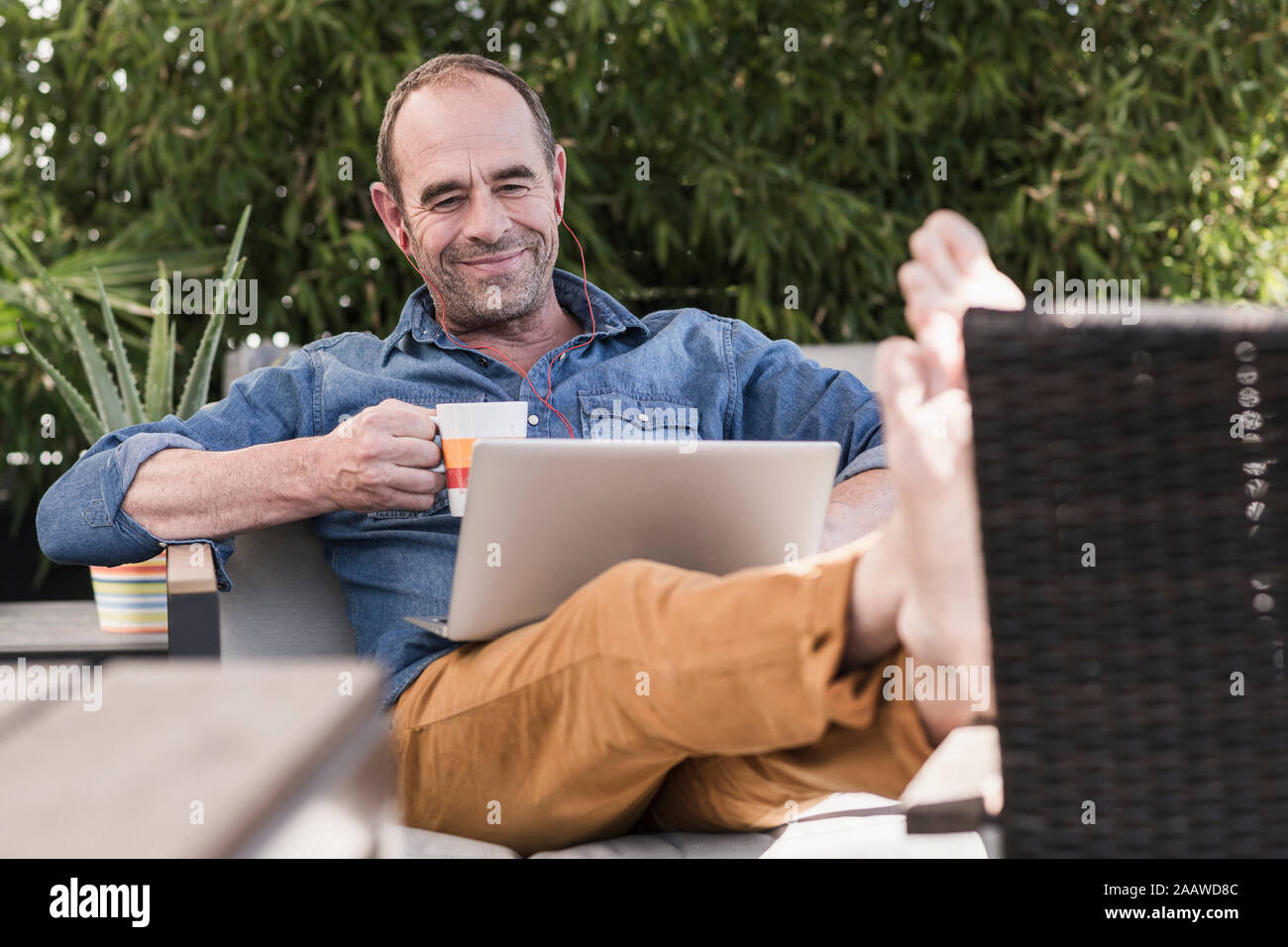 Sorridente uomo maturo seduti sulla terrazza con gli auricolari e il computer portatile Foto Stock