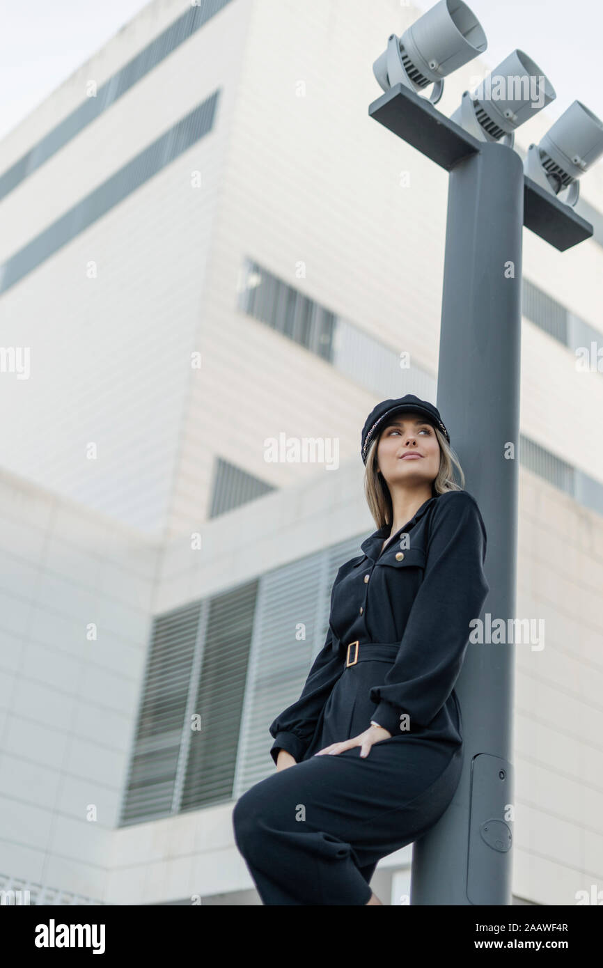 Bionda e giovane imprenditrice vestita di nero Sailor's cap e guardando lateralmente, appoggiato ad un polo della lampada a basso angolo di visione Foto Stock