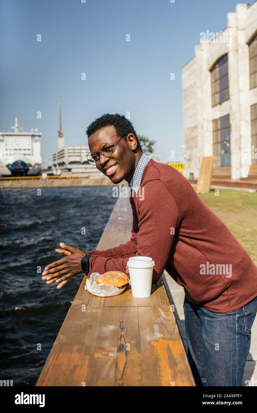 Giovane uomo che guarda il mare, mangiando hamburgerand caffè sulla ringhiera Foto Stock