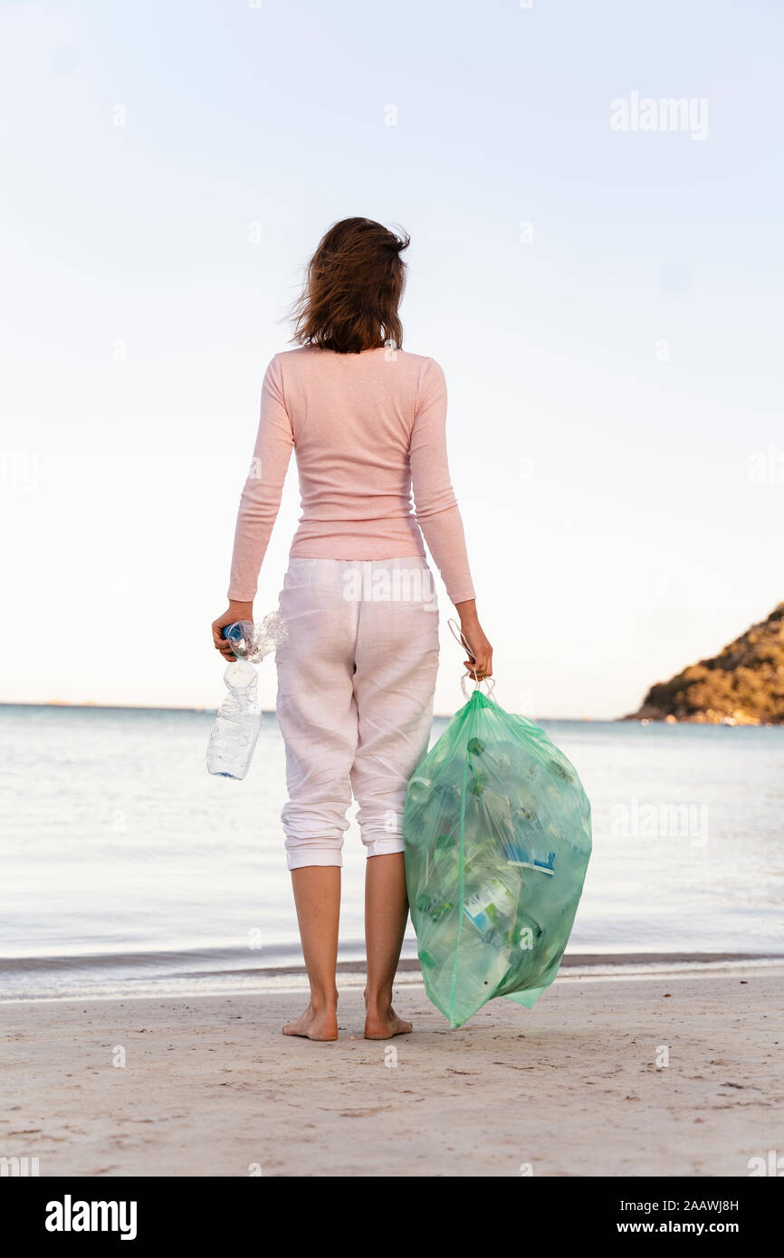 Vista posteriore della donna in piedi sulla spiaggia con bin sacchetto di raccolta le bottiglie di plastica vuote Foto Stock