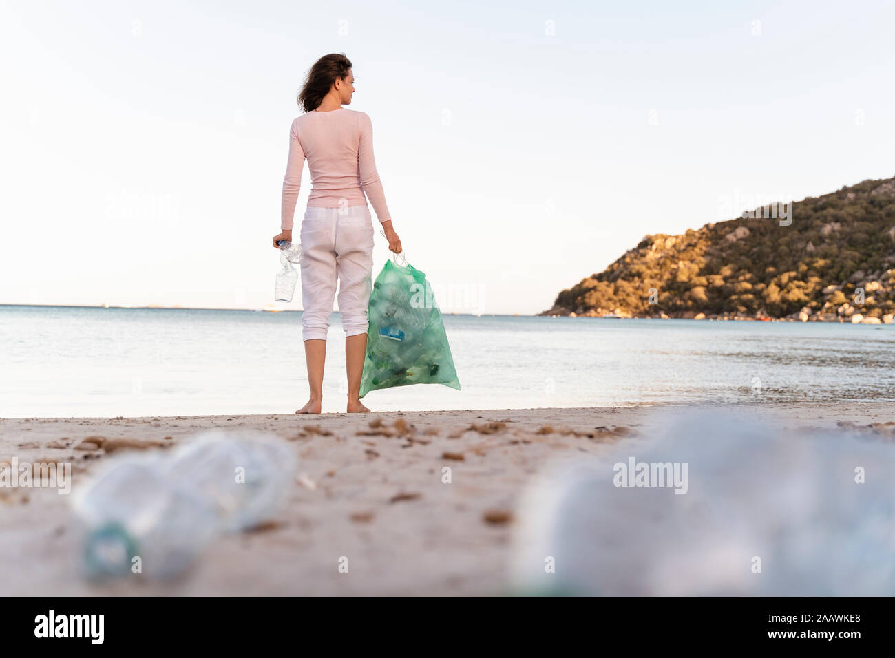 Vista posteriore della donna in piedi sulla spiaggia con bin sacchetto di raccolta le bottiglie di plastica vuote Foto Stock