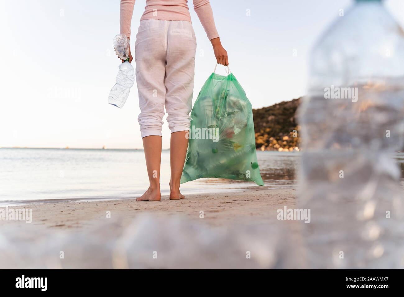 Vista posteriore della donna in piedi sulla spiaggia con bin sacchetto di raccolta le bottiglie di plastica vuote Foto Stock