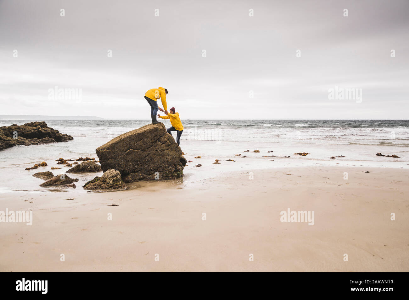 Giovane donna indossa giallo giacche pioggia e le arrampicate su roccia in spiaggia, Bretagne, Francia Foto Stock