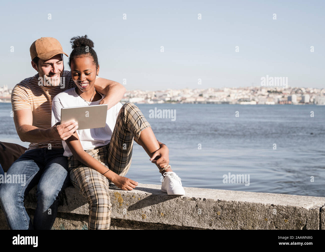 Felice coppia giovane seduto su una parete del waterfront utilizzando una compressa, Lisbona, Portogallo Foto Stock