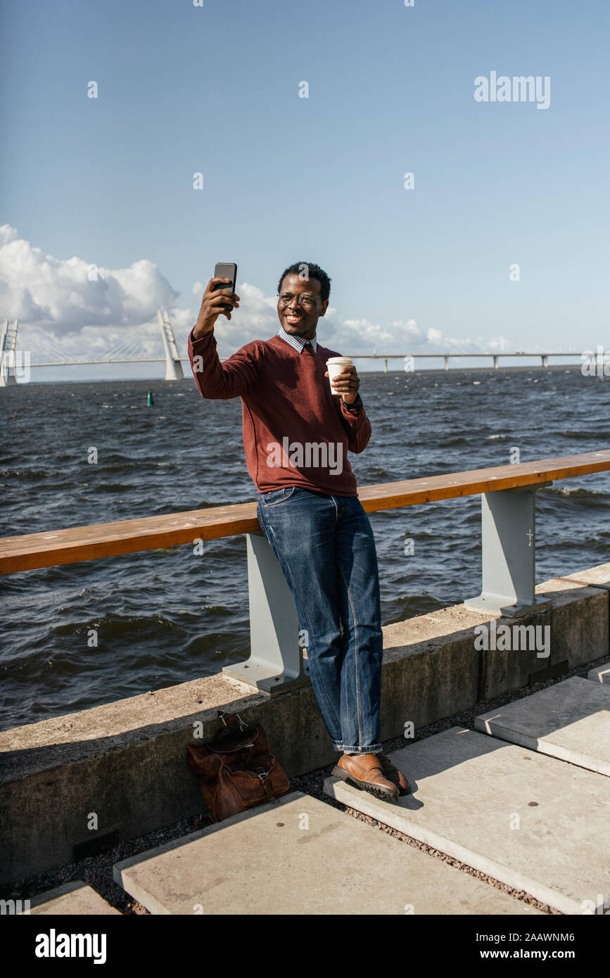 Giovane uomo prendendo selfie dello smartphone al mare, tenendo prendere il caffè Foto Stock