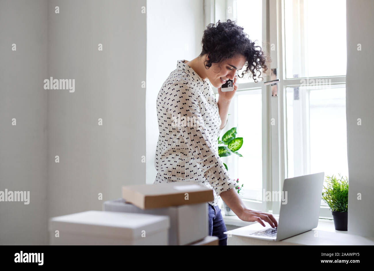 Sorridente giovane donna sul telefono cellulare utilizzando il computer portatile in ufficio in casa Foto Stock