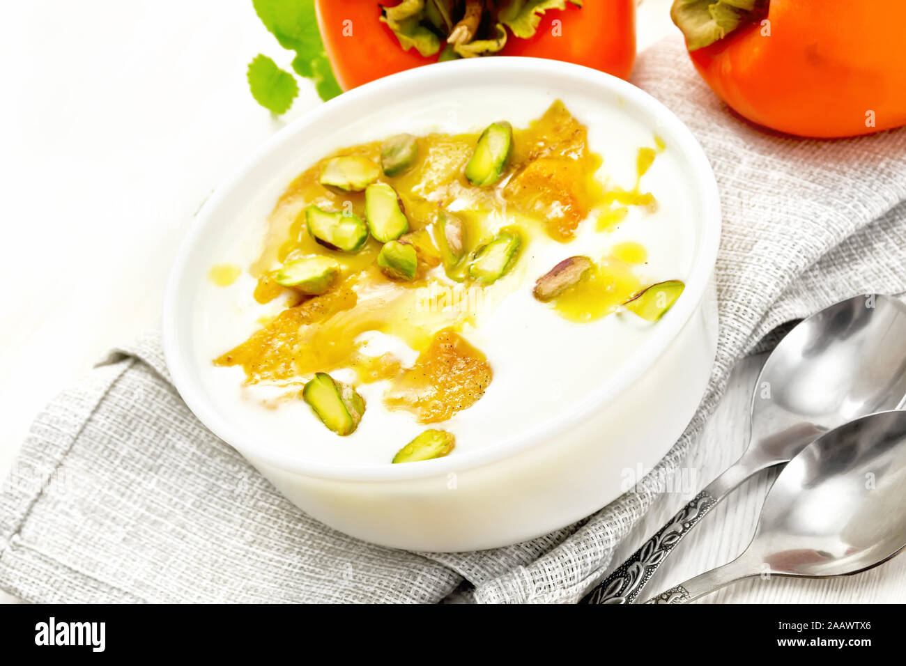 Il dessert di yogurt, persimmon e miele con vaniglia, il cardamomo e i pistacchi in una ciotola su un asciugamano, menta e cucchiai di legno sullo sfondo della scheda Foto Stock