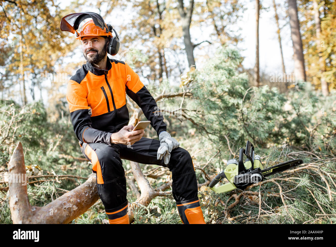 Ritratto di un professionista lumberman in indumenti da lavoro protettiva seduta con telefono cellulare sull'albero abbattuto, riposo dopo il duro lavoro della foresta Foto Stock