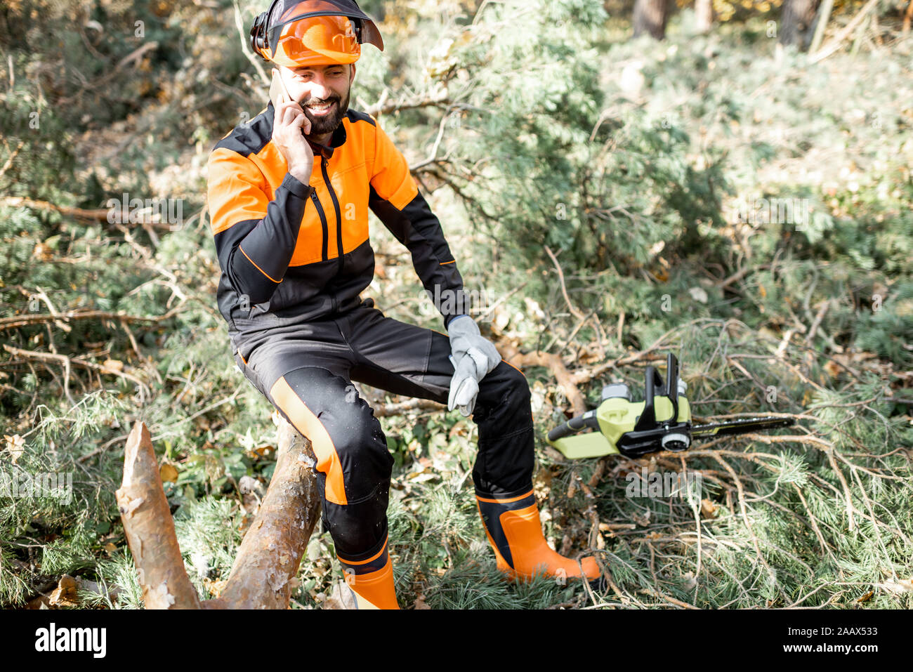 Professional lumberman in indumenti da lavoro protettiva parlando al telefono mentre è seduto sulla struttura ad albero abbattuto, riposo dopo il duro lavoro della foresta Foto Stock