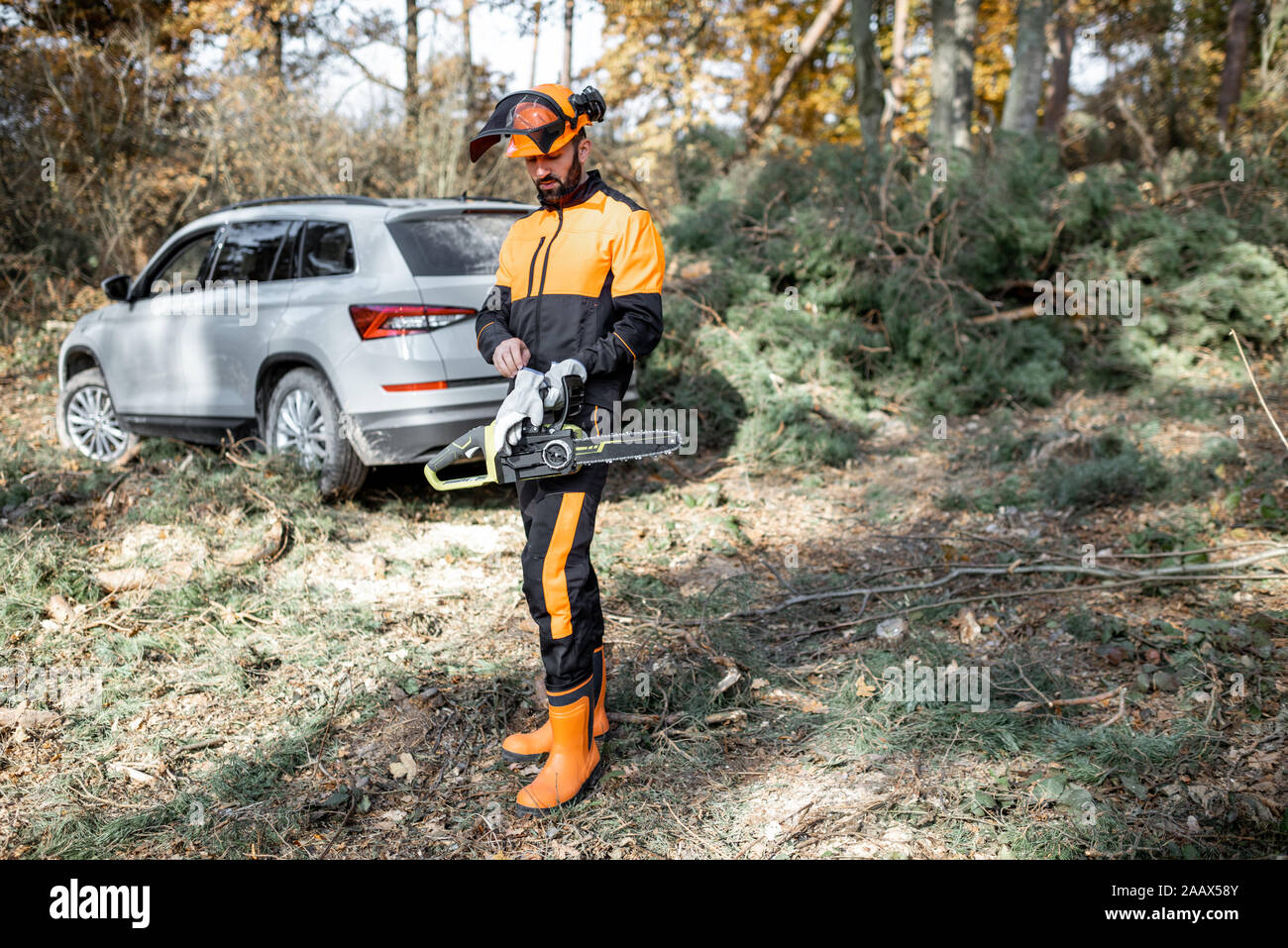Ritratto di un professionista lumberman in indumenti da lavoro protettiva registrazione con chainsaw nella foresta con auto sullo sfondo Foto Stock