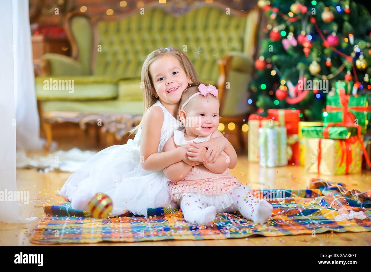 Due graziosi bambine in abiti eleganti sono seduti sul pavimento contro lo sfondo di una festosa albero di Natale. Gettare coriandoli e divertirsi. Wai Foto Stock