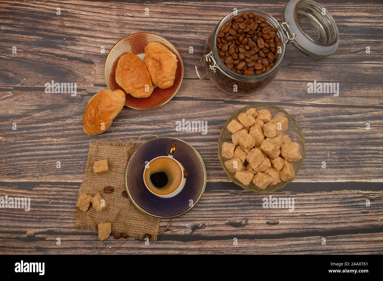 Una tazza di caffè, pezzi di zucchero di canna in una zuccheriera, i chicchi di caffè in un vasetto di vetro e croissant su uno sfondo di legno Foto Stock