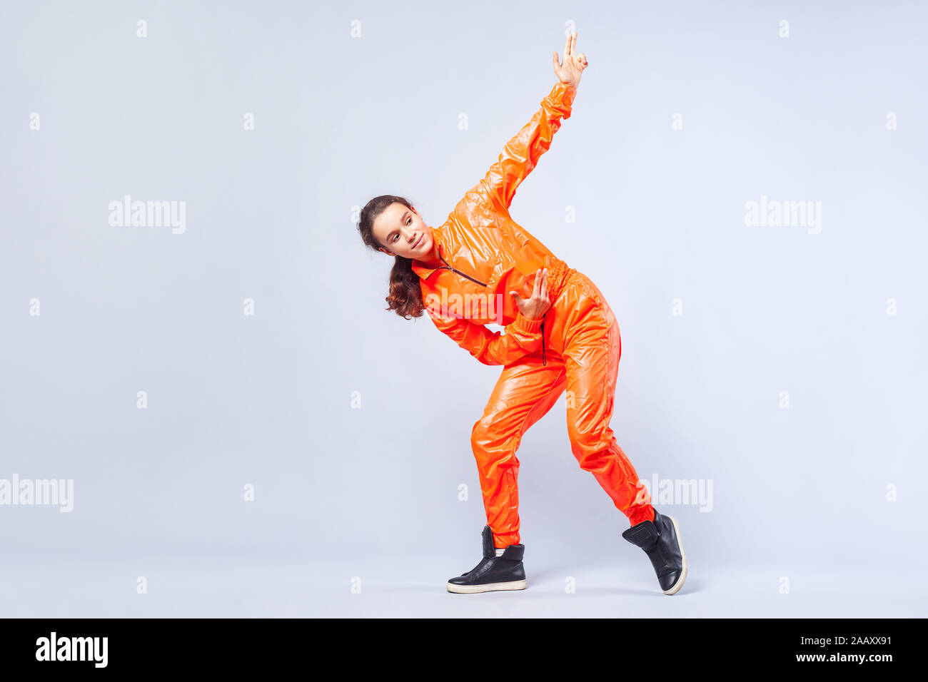 A piena lunghezza Ritratto di energetico positivo teen ragazza con brunette capelli luminosi in tuta arancione danza hip-hop e rivolta verso l'alto con un dito la pistola Foto Stock