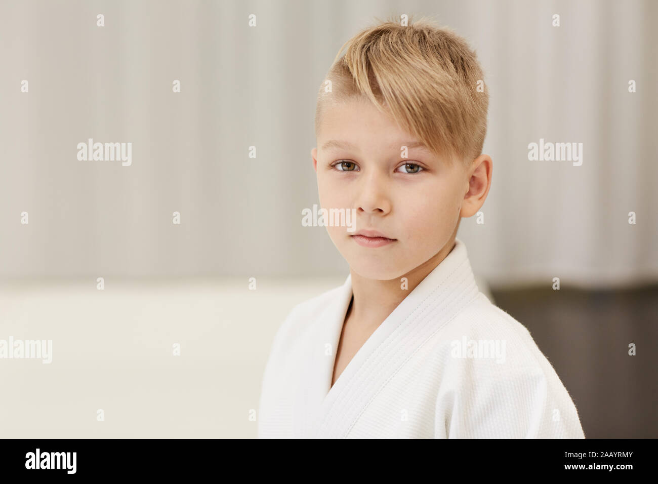 Ritratto del ragazzino con i capelli biondi e in kimono guardando la telecamera mentre sta in piedi in palestra Foto Stock