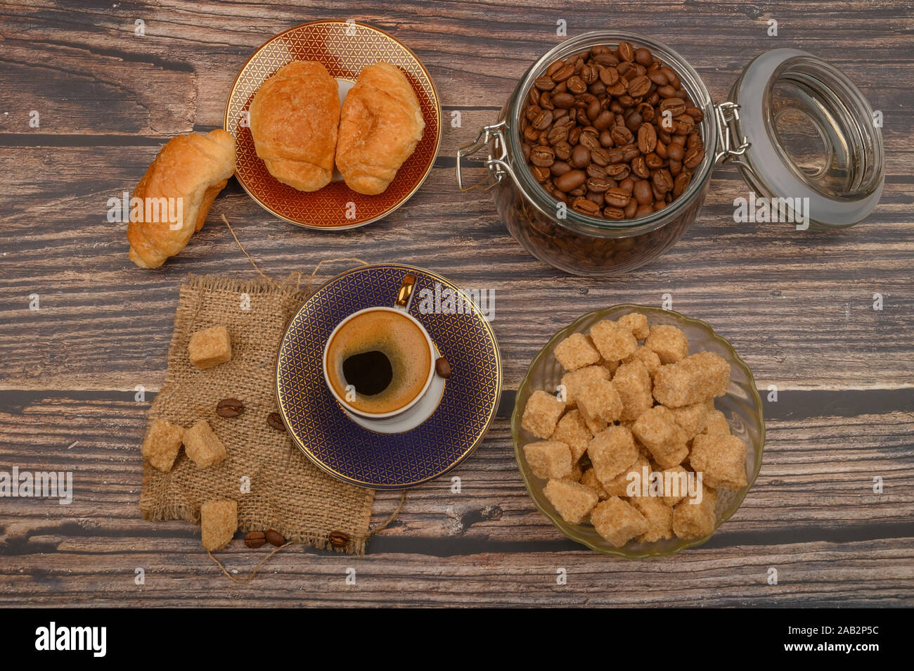 Una tazza di caffè, pezzi di zucchero di canna in una zuccheriera, i chicchi di caffè in un vasetto di vetro e croissant su uno sfondo di legno Foto Stock