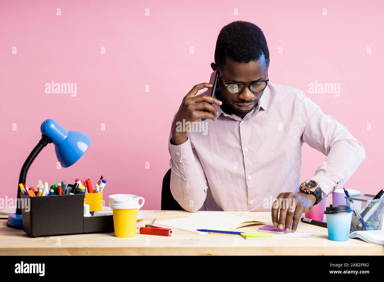 Pragmatico barba lunga african american uomo che indossa gli occhiali, camicia a righe, parlando con il partner aziendale sullo smartphone, seduti alla scrivania circondata wit Foto Stock