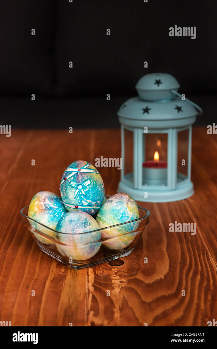 Decorazione di Pasqua - blu, verniciato colorato uova con pietre preziose in una ciotola di vetro e una blu lanterna a candela su una tavola di legno Foto Stock