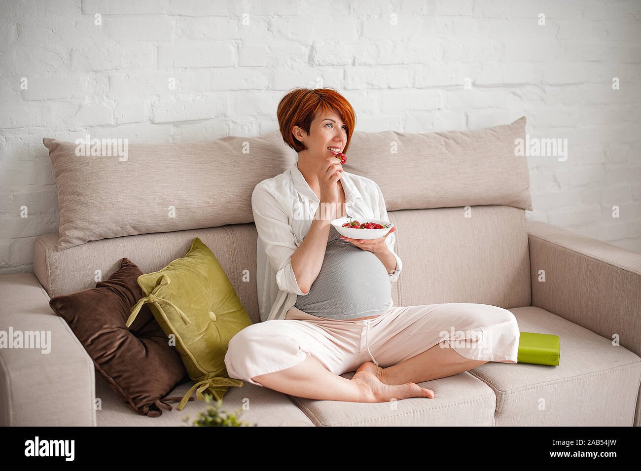 Felice donna incinta di mangiare fragole su un divano a casa Foto Stock