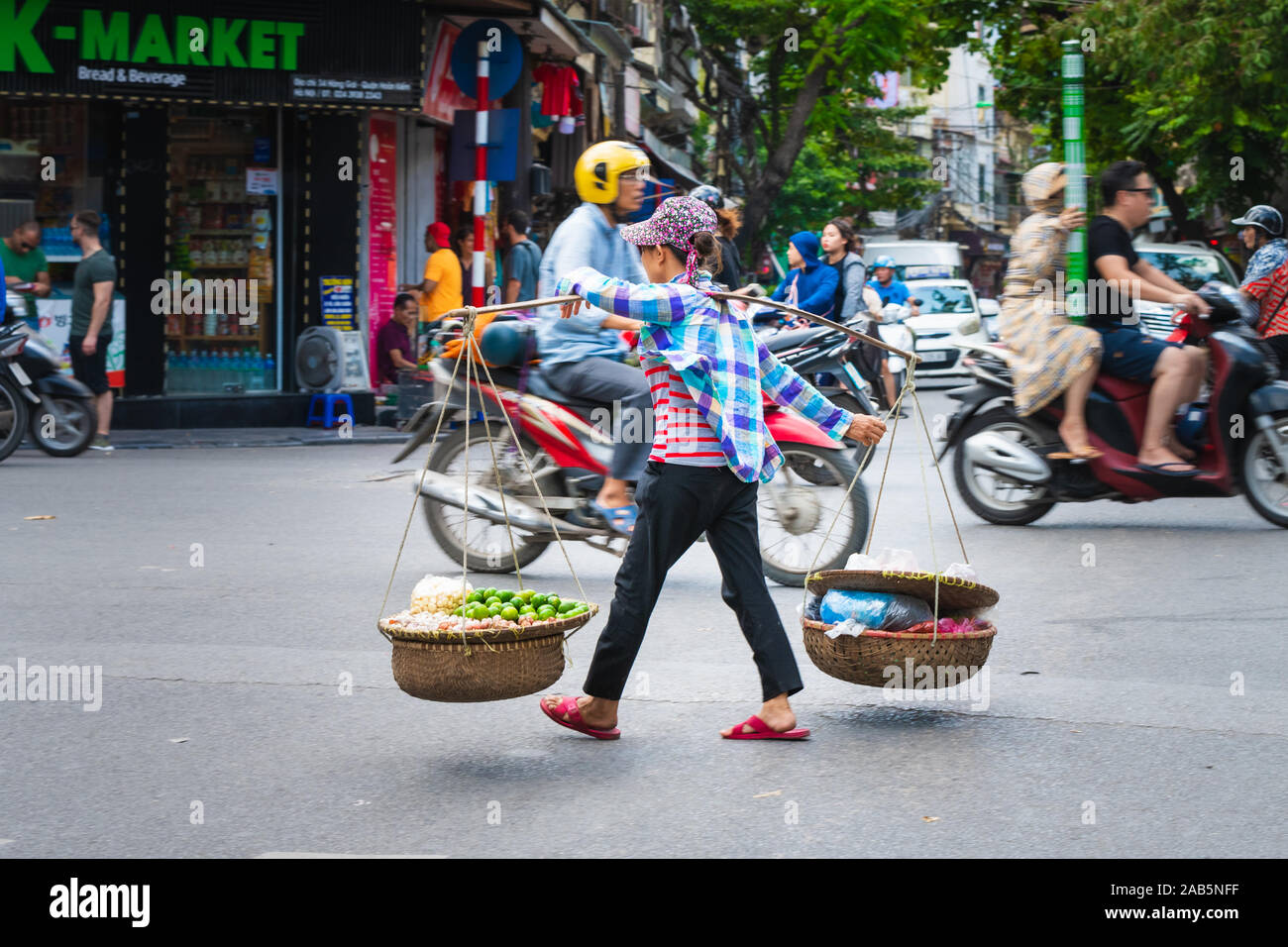 Hanoi, Vietnam - 11 Ottobre 2019: donna asiatica con una portante in legno pole di trasportare merci attraverso una strada trafficata di Hanoi Asia Foto Stock