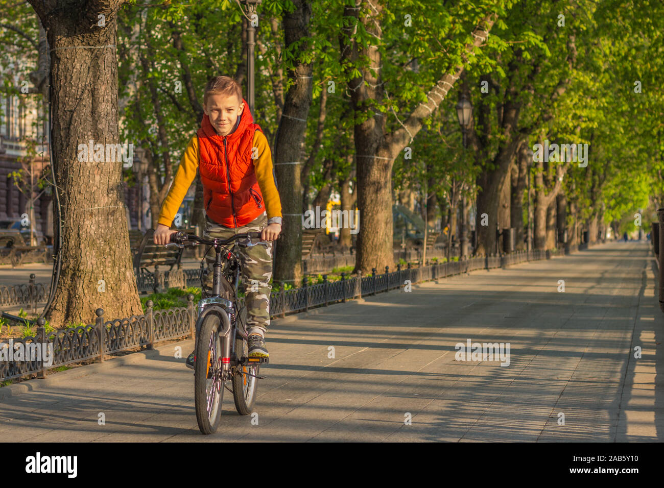 Un ragazzo di 8 anni in un giubbotto arancione corse in bicicletta in un parco della città su una soleggiata mattina di primavera Foto Stock
