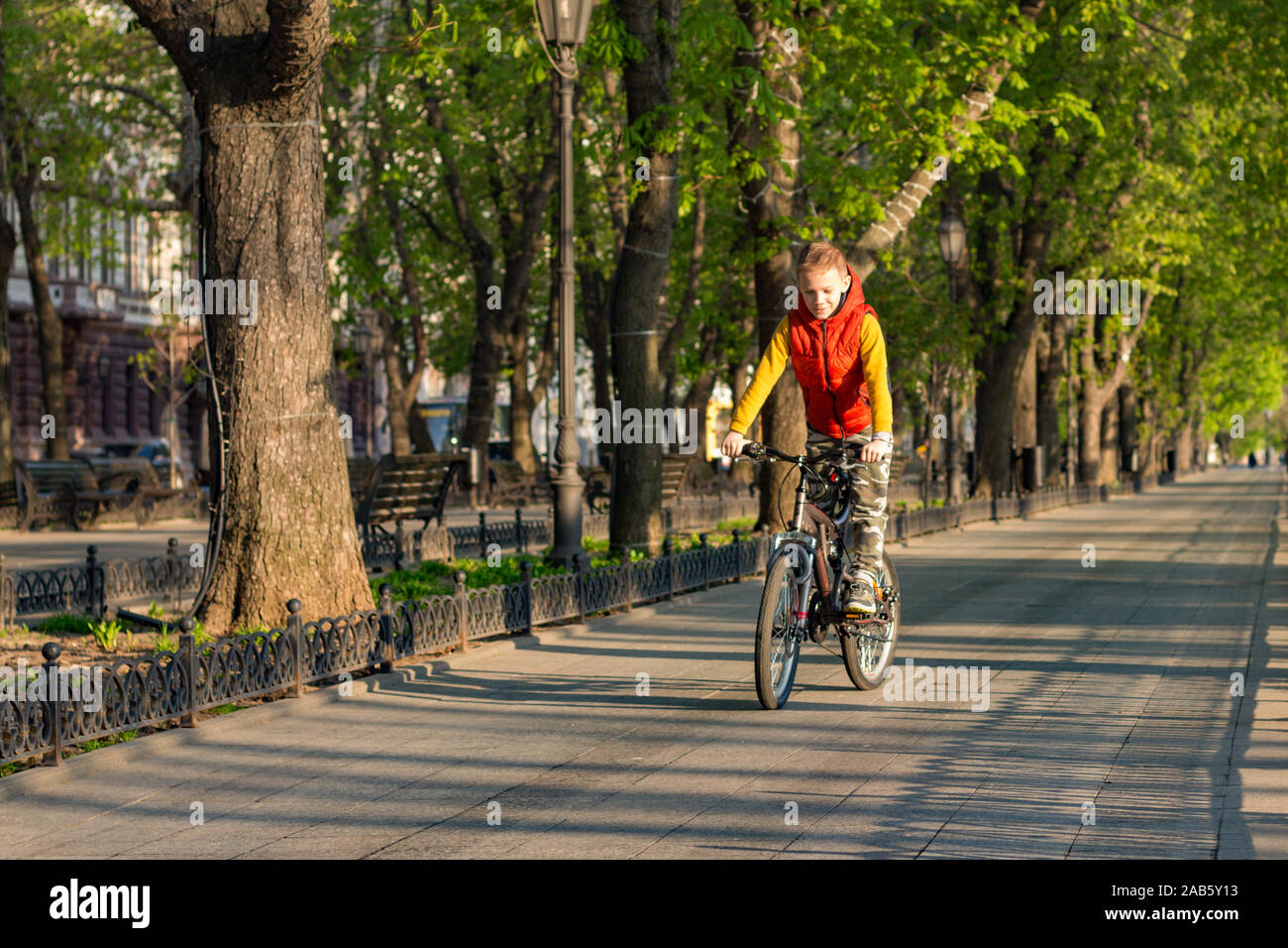 Un ragazzo di 8 anni in un giubbotto arancione corse in bicicletta in un parco della città su una soleggiata mattina di primavera Foto Stock