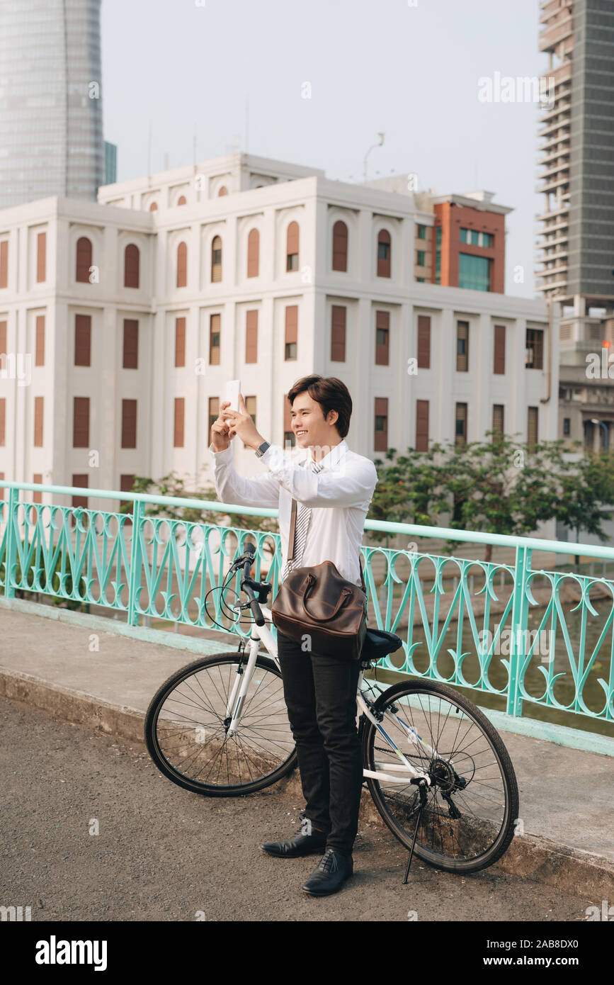 Giovane uomo di scattare una foto con il suo smart phone mentre in piedi sulla strada di città con la sua bicicletta accanto a lui Foto Stock