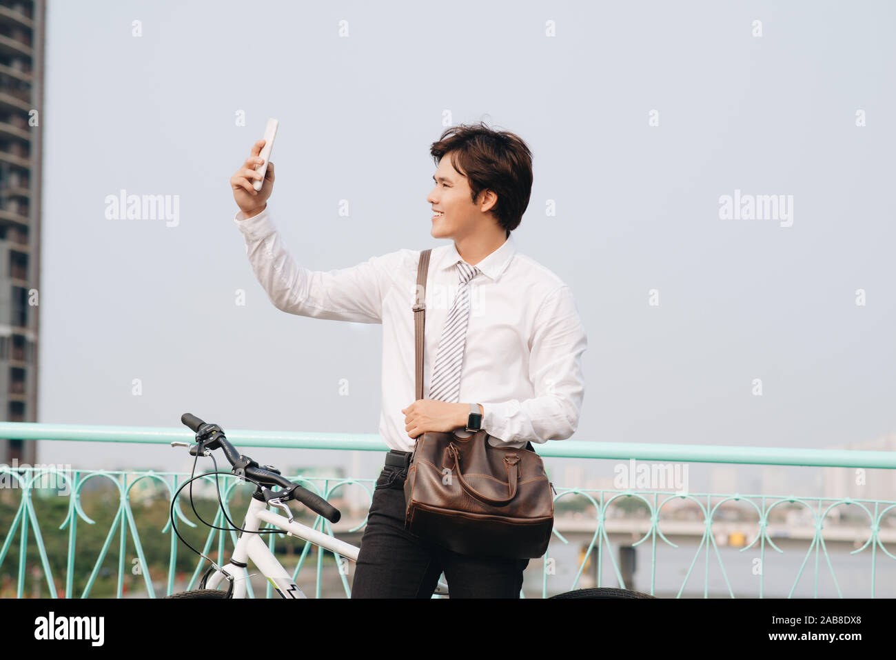 Giovane uomo di scattare una foto con il suo smart phone mentre in piedi sulla strada di città con la sua bicicletta accanto a lui Foto Stock