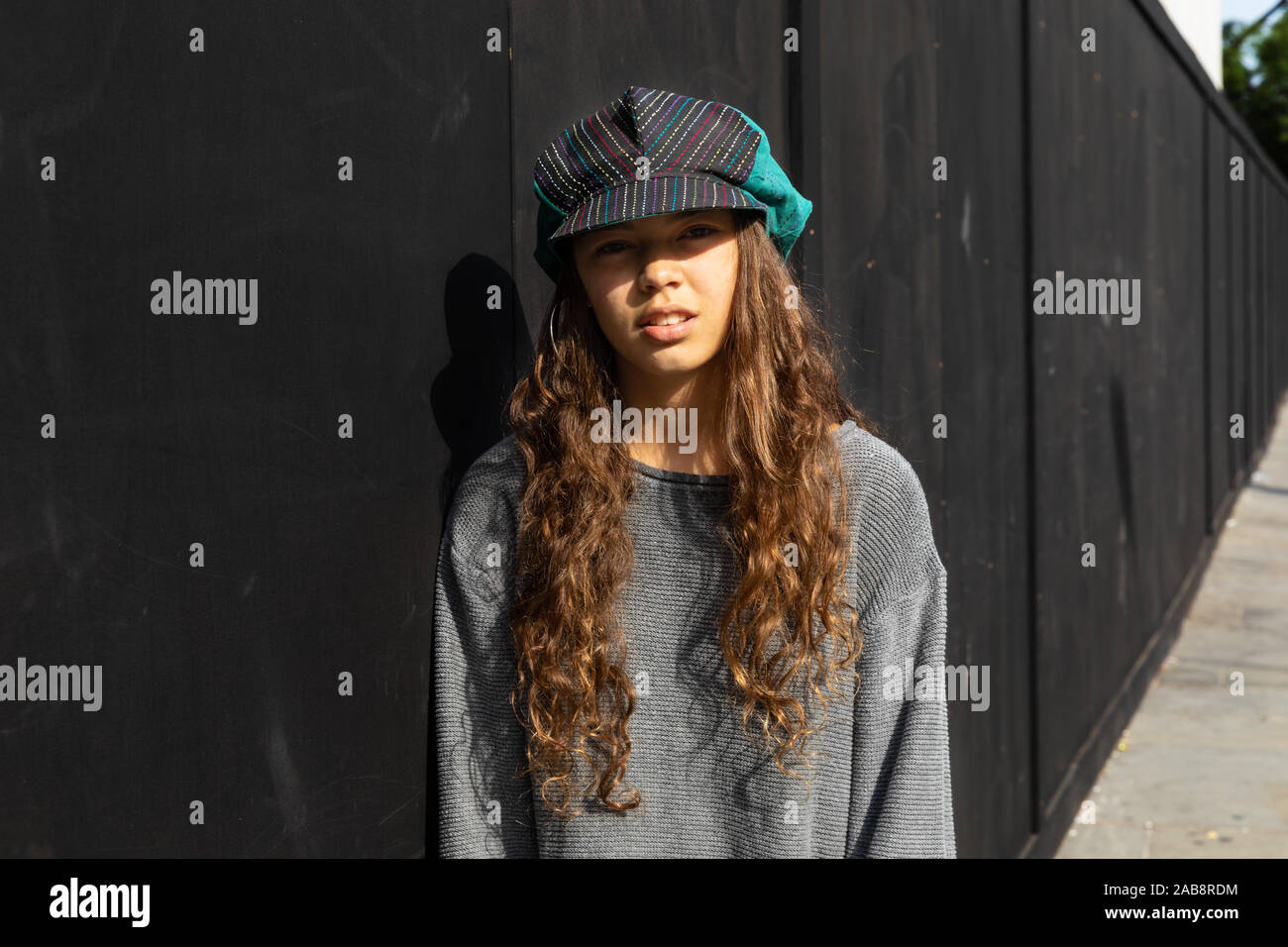 Emotionless ragazza con colorati hat e capelli lunghi marrone. Contesto urbano e sfondo nero. Foto Stock