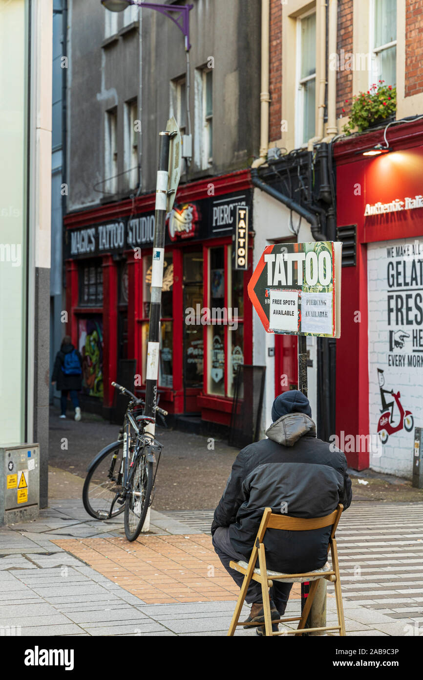 Scheda sandwich pubblicità uomo seduto con annunci di salotto tattoo nella città di Cork, Irlanda Foto Stock