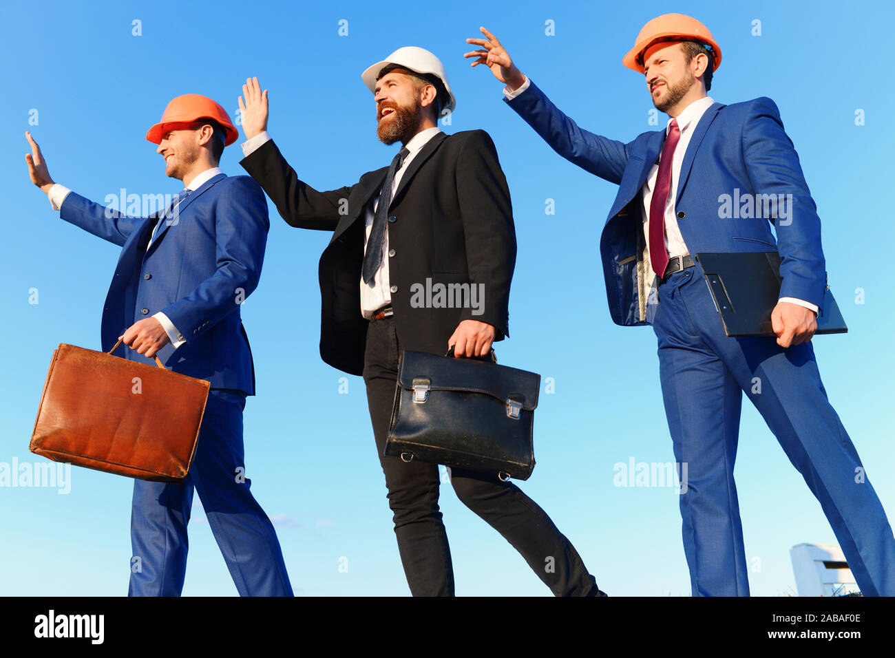 Costruttori a piedi e wave a qualcuno sul cielo blu sullo sfondo. Coworking e costruzione concetto di business. Gli architetti con volti felici in tute e caschi. Gli ingegneri della cartella in attesa, nero e marrone casi Foto Stock