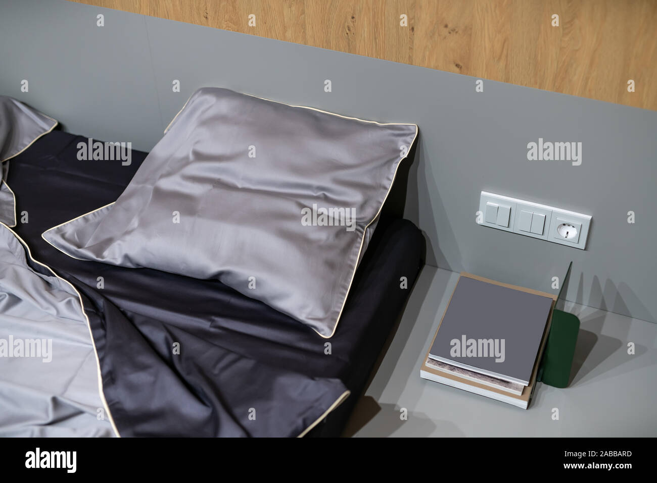 Splendido posto letto con lenzuola di seta sul pavimento grigio nel luminoso interno con pareti in legno. Accanto ad essa ci sono libri e una presa di alimentazione Foto Stock