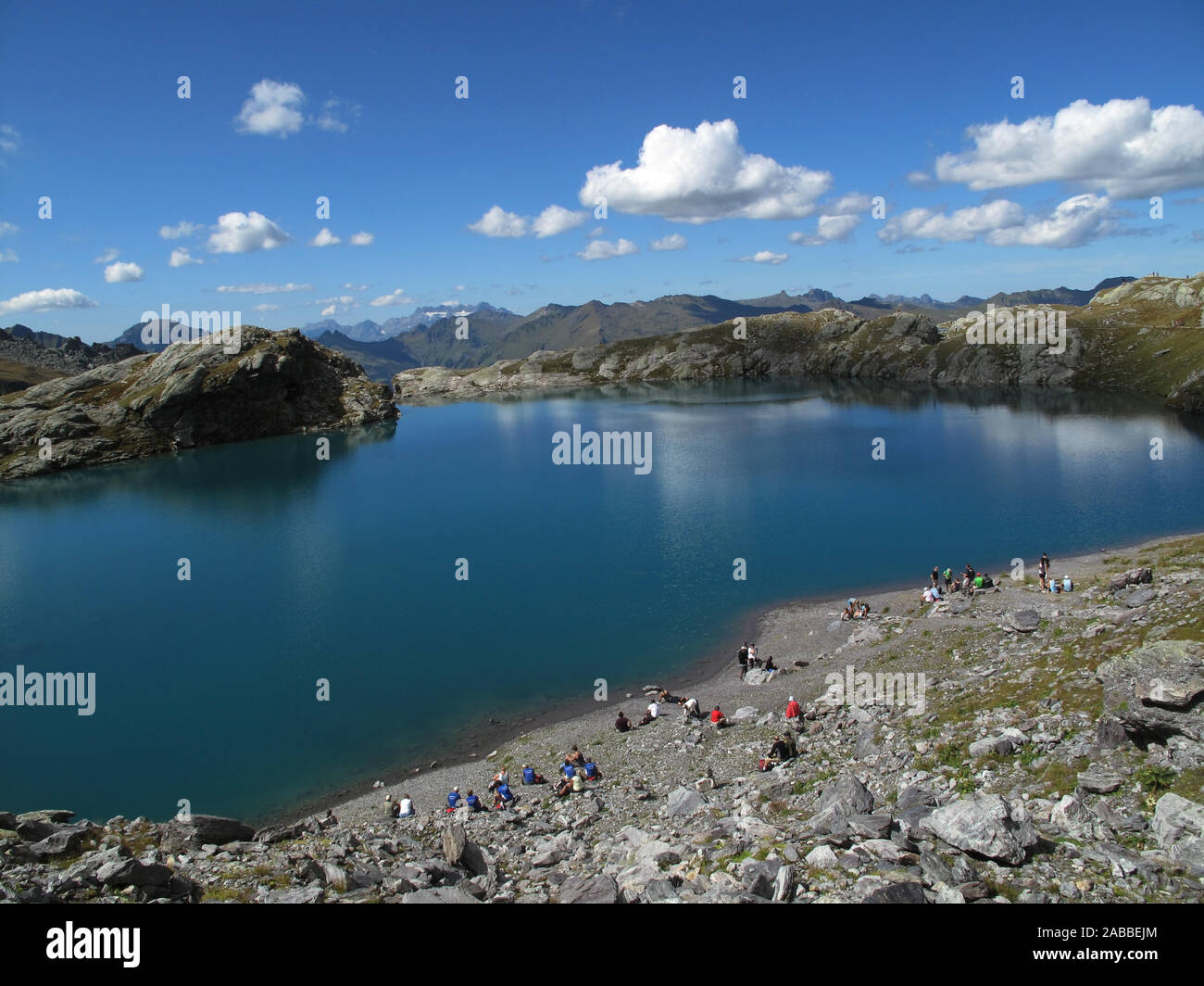 Alpi svizzere. Vicino al cielo accanto al profondo blu del lago di montagna con riflessi in acqua fluttuante molti escursionisti di riposo. Dietro di loro rocce un blu cielo nuvoloso Foto Stock