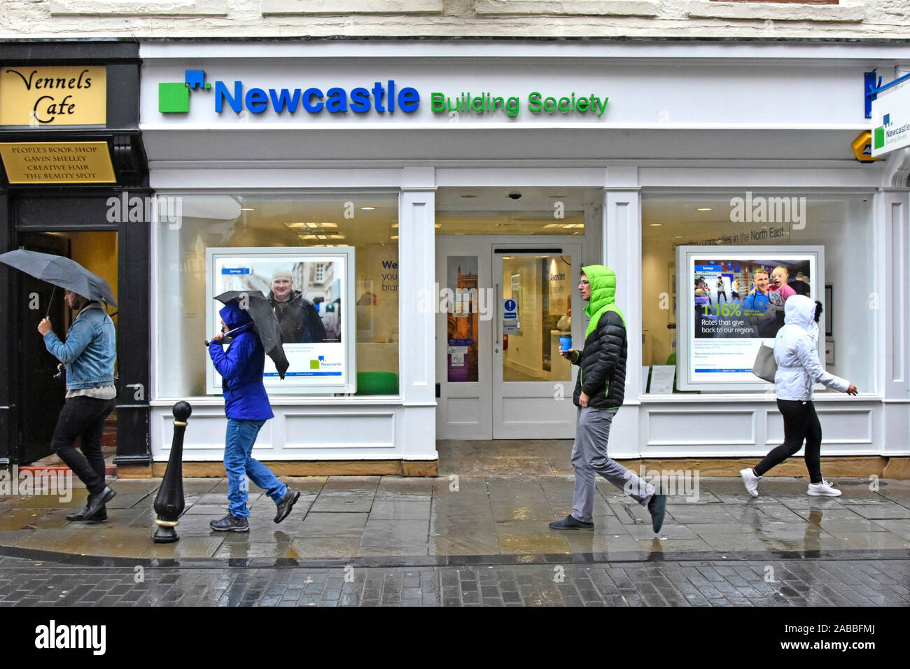 Scena di strada & studio candidi persone vestite per affrontare una giornata di pioggia camminando lungo il marciapiede bagnato alcuni ombrelloni nella strada dello shopping di Durham Regno Unito Inghilterra Foto Stock